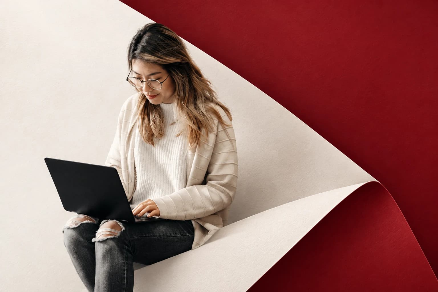 Photo illustration of a woman using a laptop, composed with an oversized cream paper fold against burgundy.